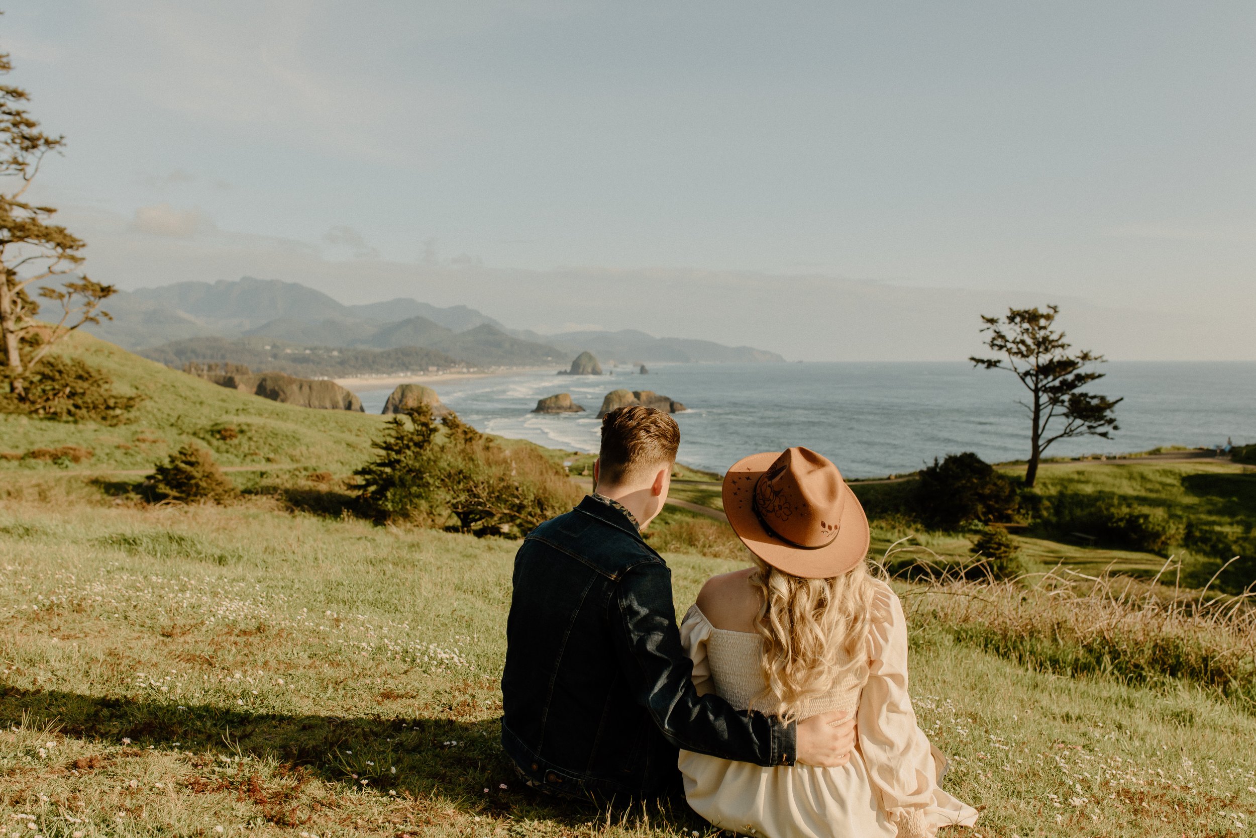 man holding woman while both are sitting down on grass overlooking entire ocean and beach in Oregon, Oregon film photography
