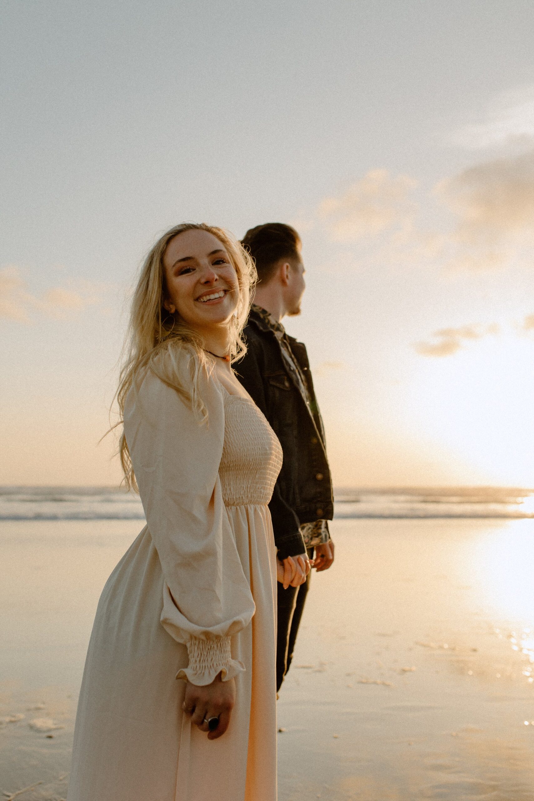  woman smiling at camera while holding mans hand and standing on beach near ocean 