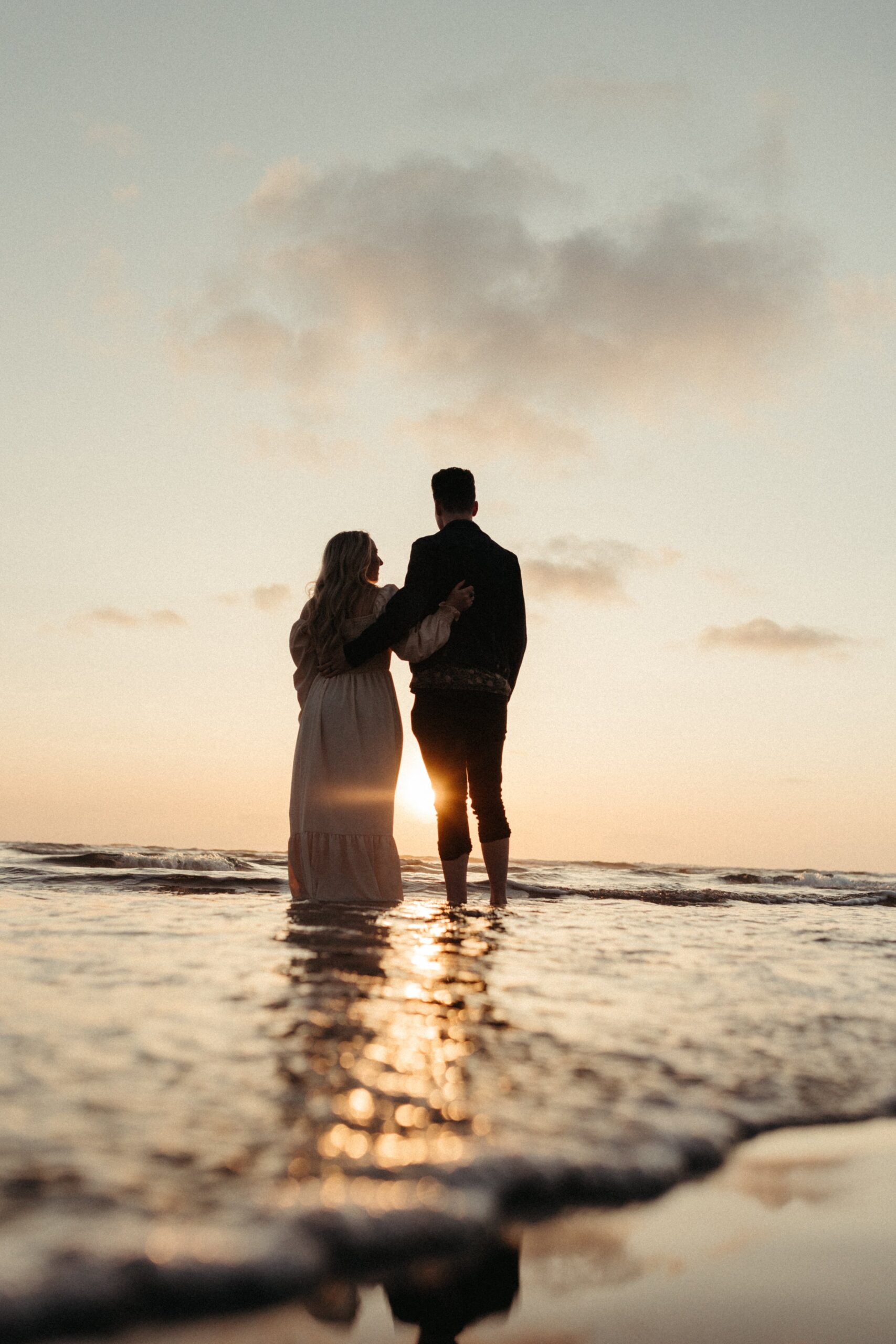 man and woman holding hands while standing on the beach during sunset