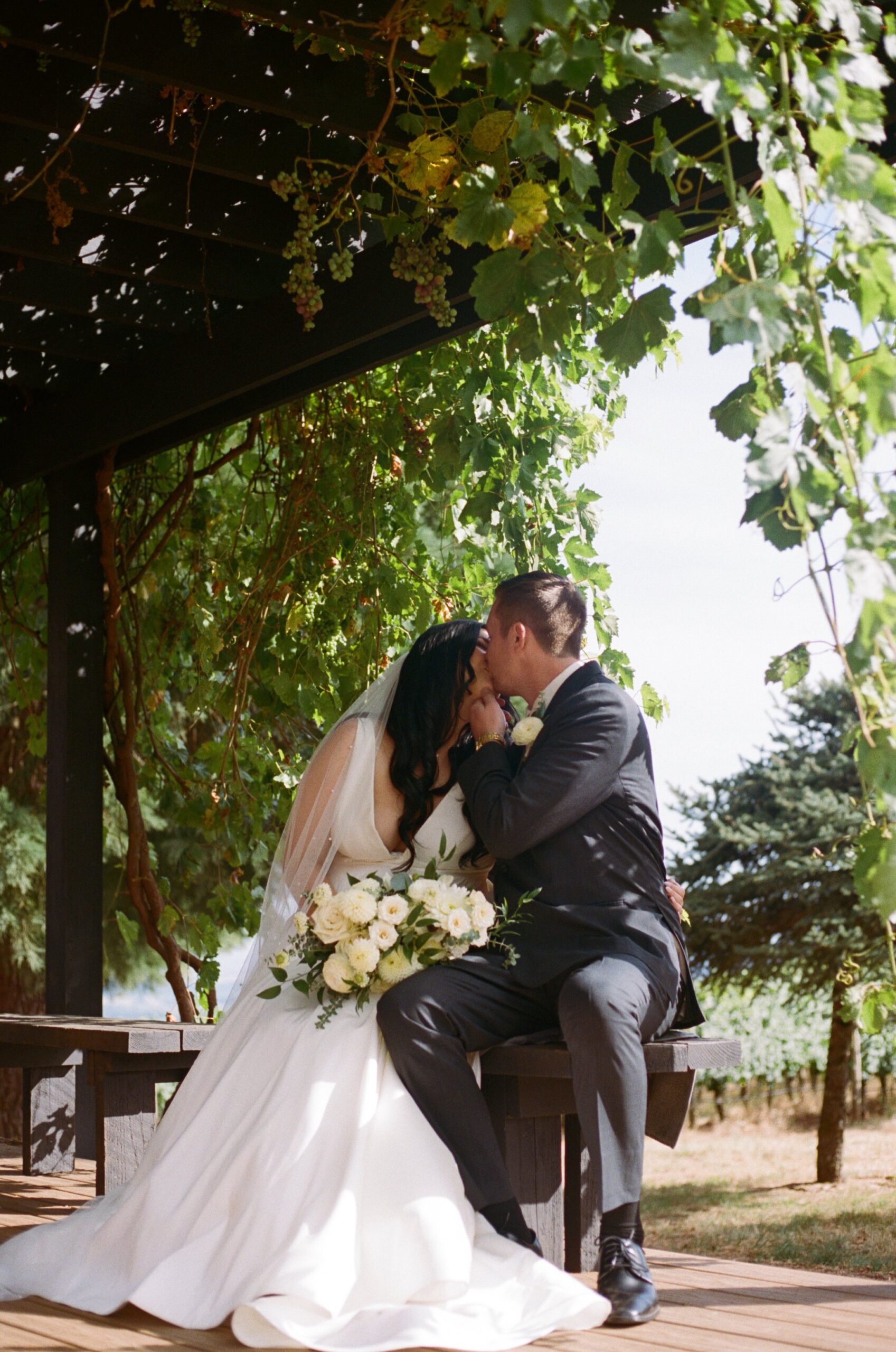 bride and groom kissing under grape vines while bride is holding bouquet of flowers