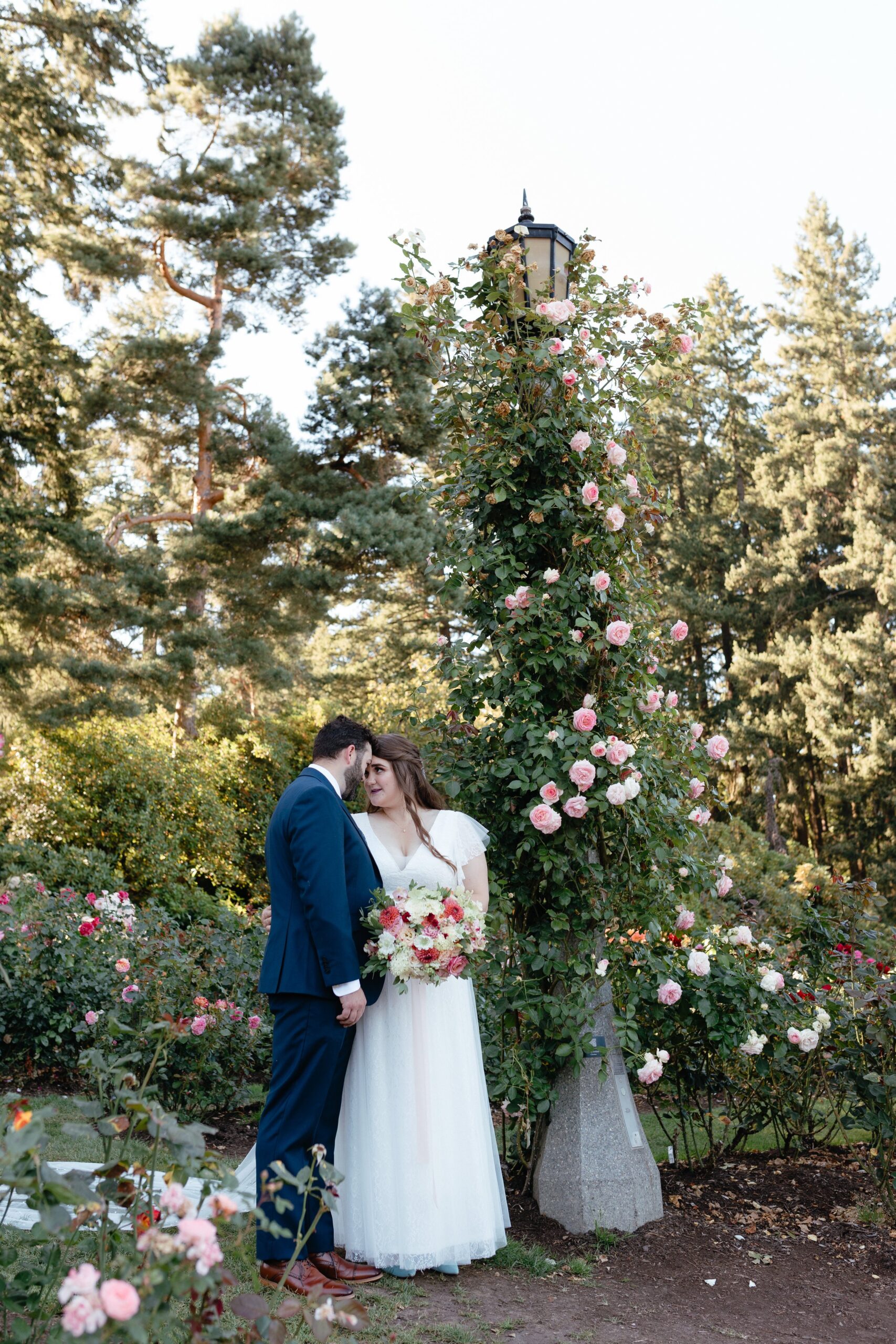  bride in white dress holding pink bouquet, groom in navy blue suit, holding hands, kissing, Oregon elopement, portland international rose garden 