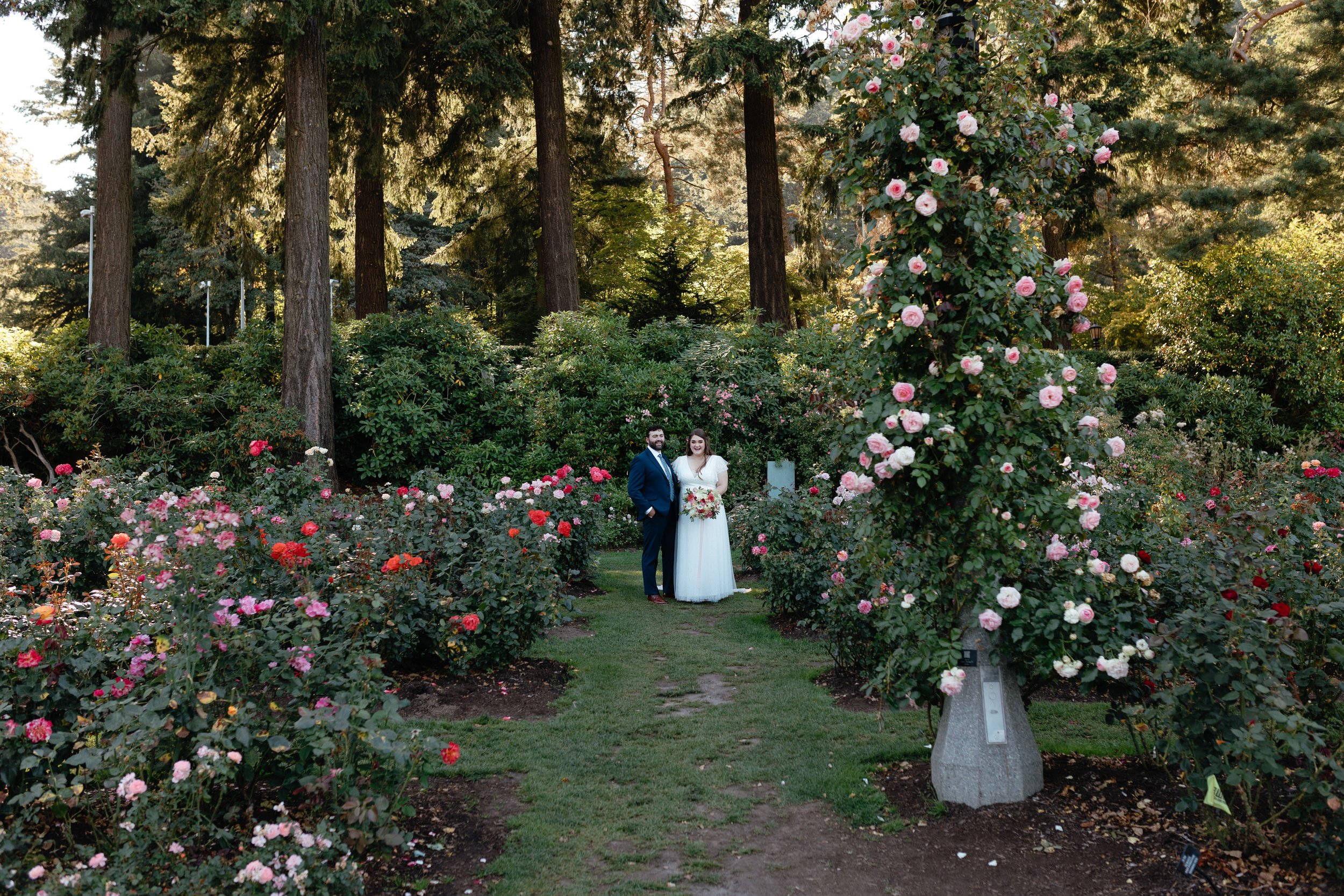  bride in white dress holding pink bouquet, groom in navy blue suit, holding hands, kissing, Oregon elopement, portland international rose garden 
